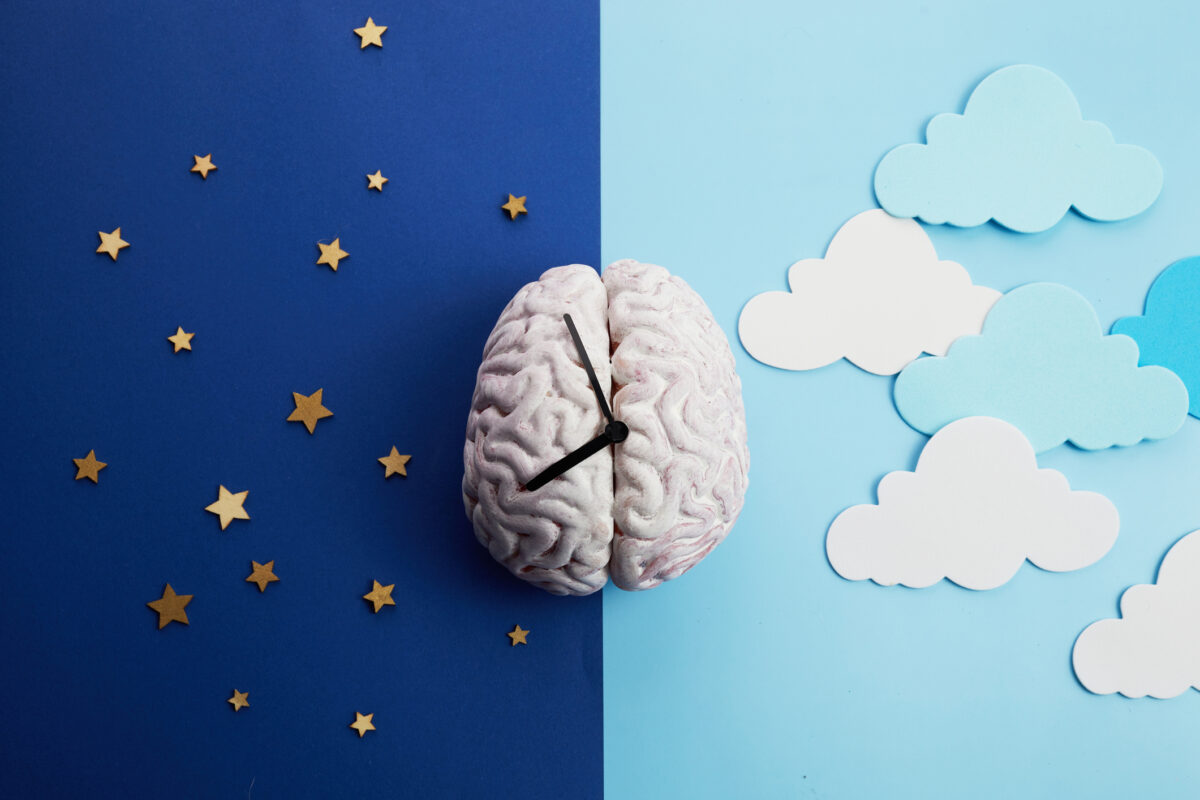 Image is of a brain with a clock face on it. The left side of the image is dark blue with gold stars indicating nighttime; the right side of the image is light blue with clouds, indicating daytime.