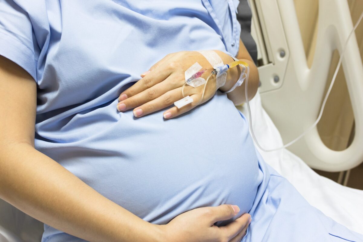 A pregnant woman wearing a hospital gown and with an IV in her hand cradles her belly