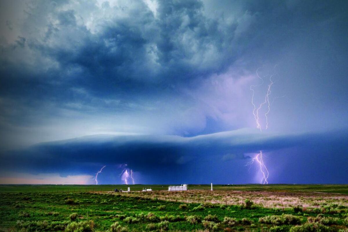 This image shows a stormy sky over a vast, open field. Dark clouds loom overhead, with multiple lightning bolts striking in the distance. A few small structures sit on the horizon, contrasting with the turbulent sky.