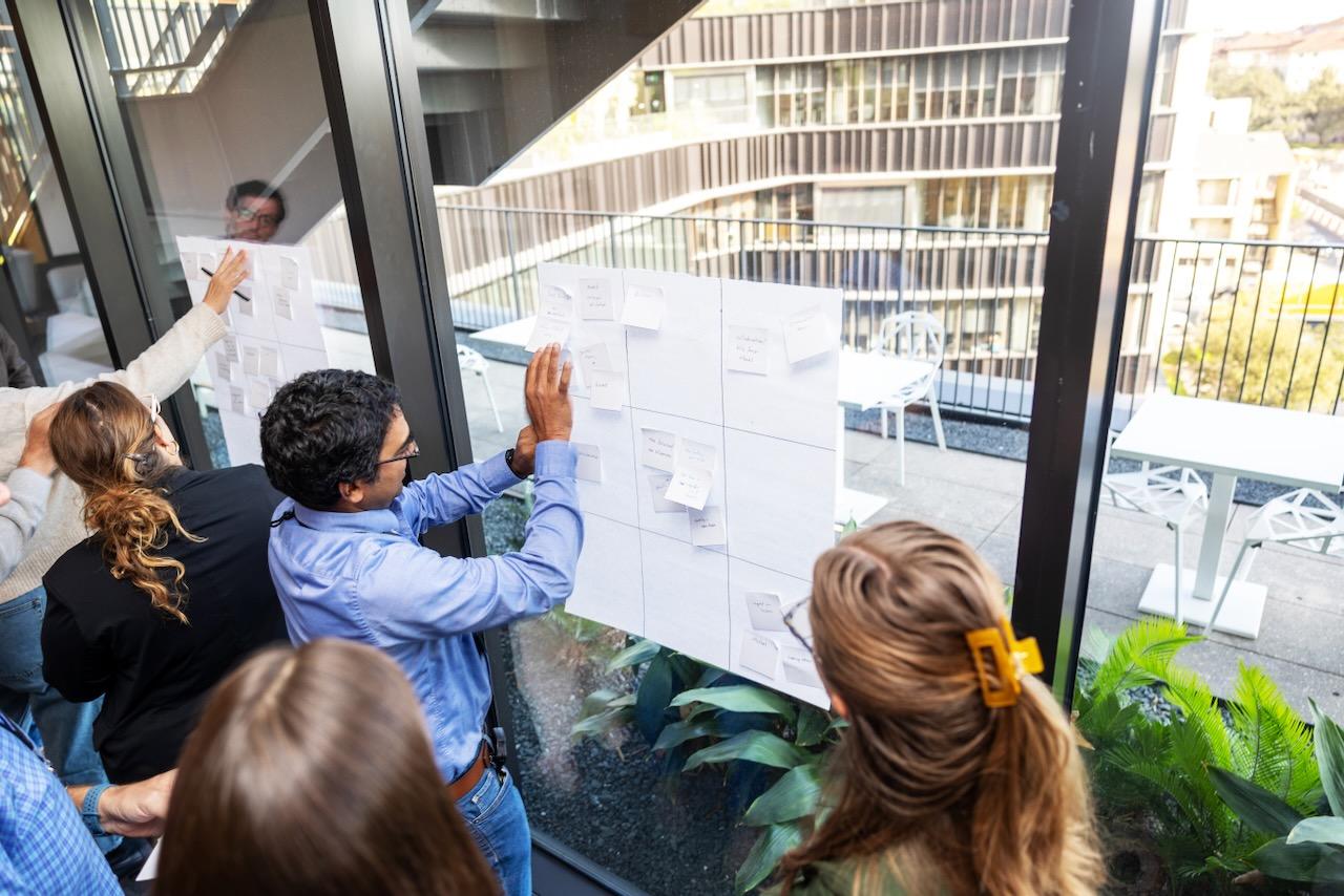 A UT faculty memer is posting notes on a poster-sized piece of paper hanging on a window. He is surrounded by fellow UT faculty members participating in APX