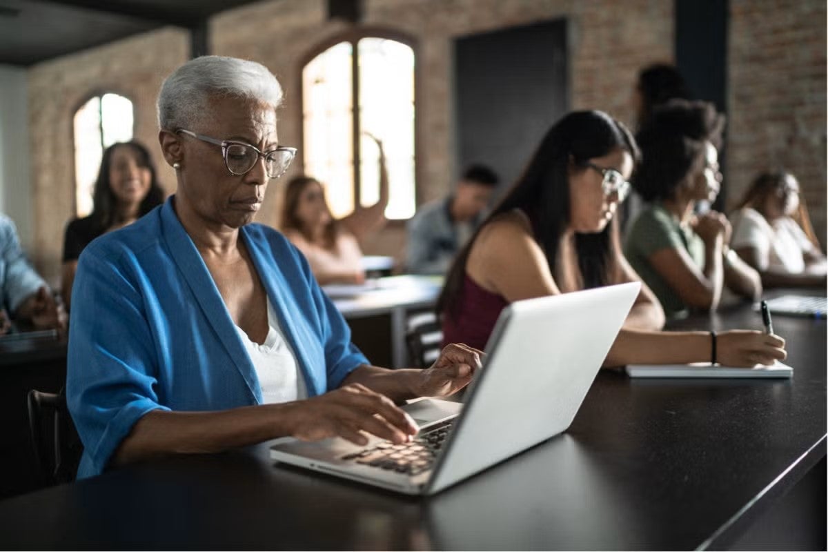 The image shows a diverse group of people seated in a classroom setting. The focus is on an older individual with short white hair, wearing glasses and a blue cardigan, typing on a laptop. Other individuals are seen in the background, some taking notes and others using electronic devices. The room has large windows and exposed brick walls.