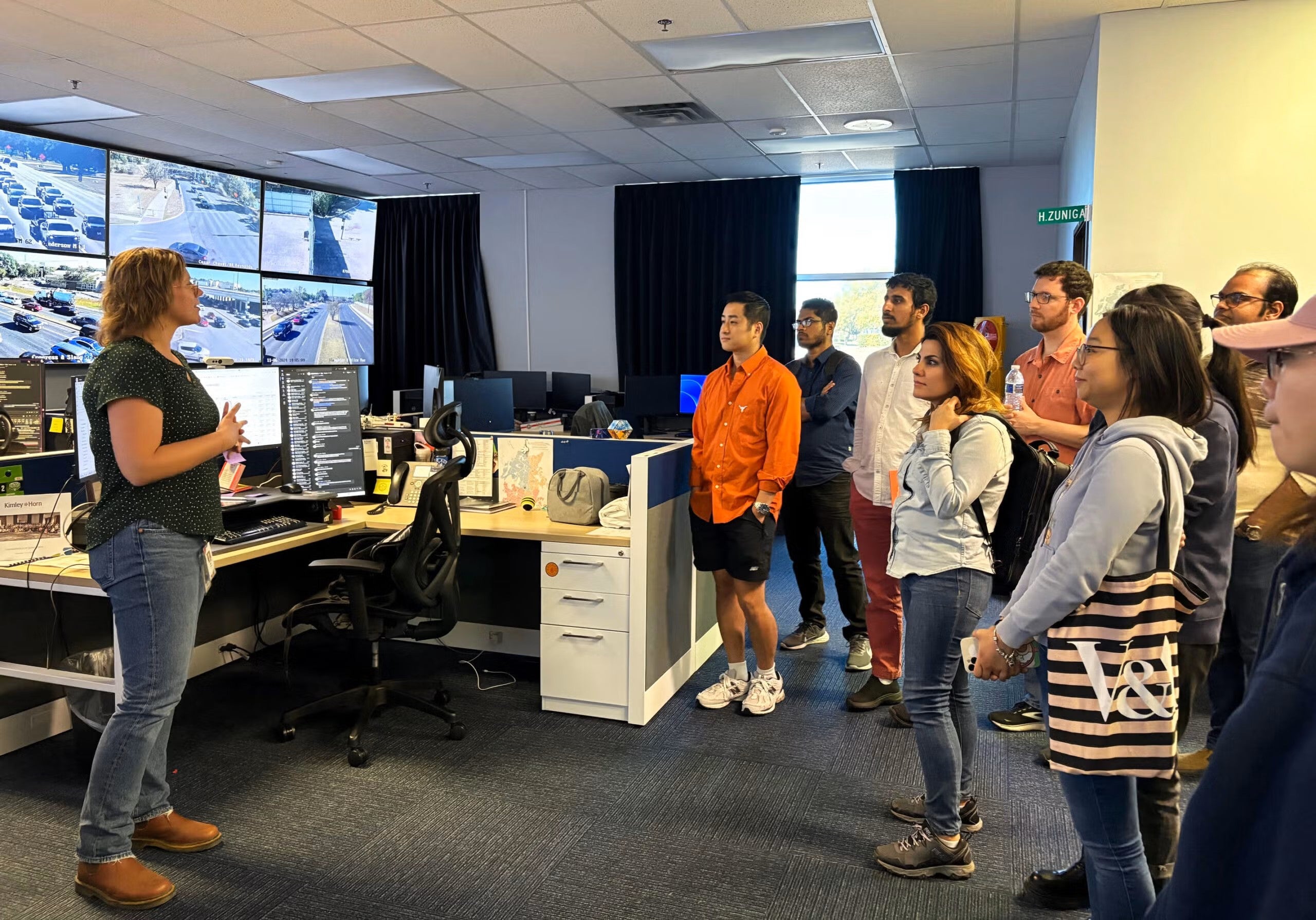 A woman standing in front of several screens speaks to a group of people.
