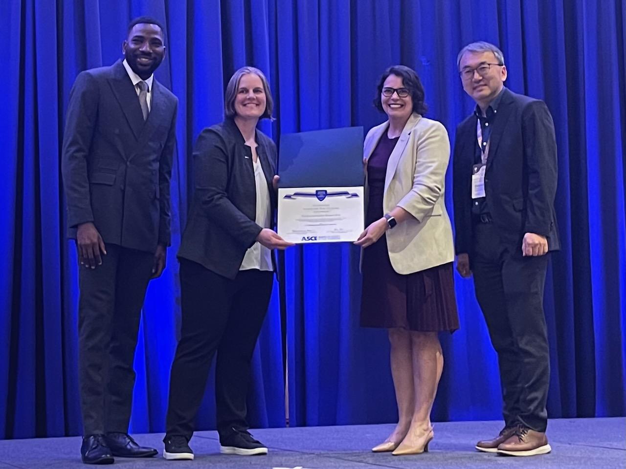 Leite stands on stage with three others, holding the certificate presented to her for winning the 2026 Peurifoy Construction Research Award from the American Society of Civil Engineers.