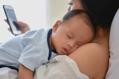 Baby sleeps on mother's shoulder as she looks at cell phone